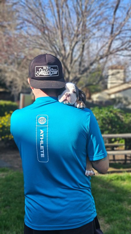 A person in a blue shirt holds a small dog against their shoulder outdoors.