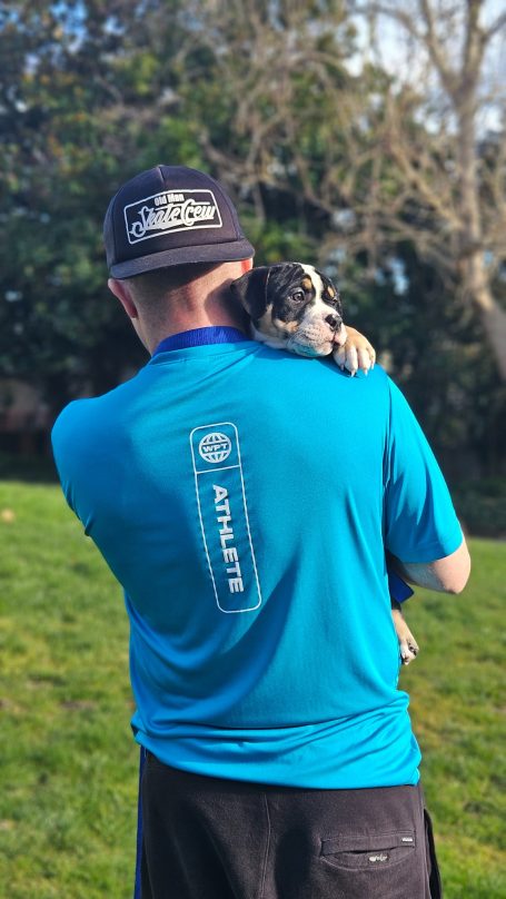 A person in a blue shirt holds a small dog against their shoulder outdoors.